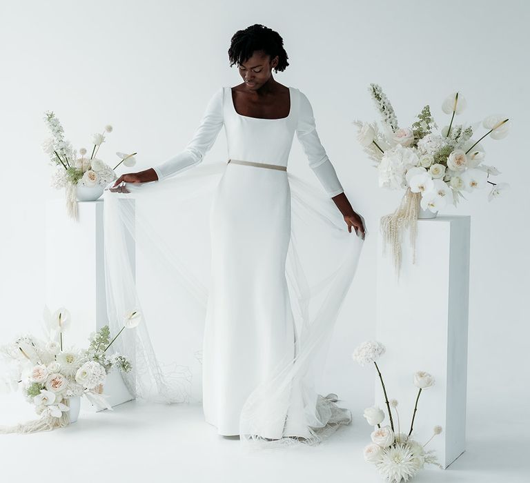 Black bride in a fitted wedding dress with long sleeves wearing a detachable embroidered tulle skirt standing in the centre of her all white wedding flower arrangements 