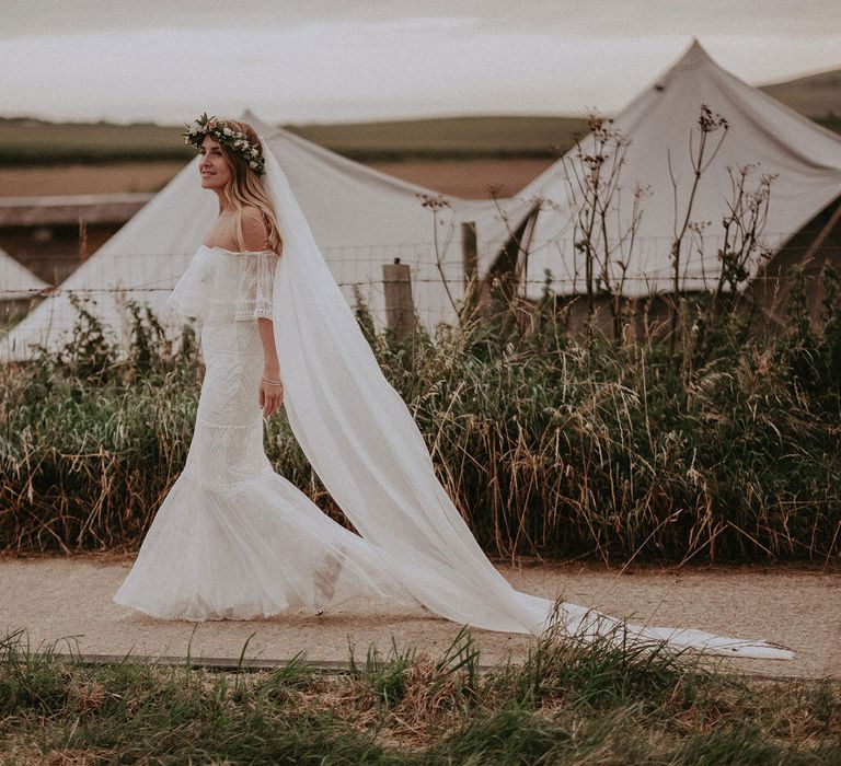 Bride in Grace Loves Lace bardot wedding dress, veil and flower crown walks along path by white tipis at Isle of Wight wedding with macrame wedding decor