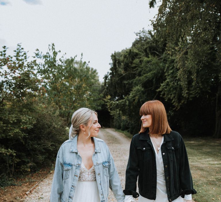 Two brides walk hand in hand on gravel path, one bride wears a leather jacket and the other wears a denim jacket