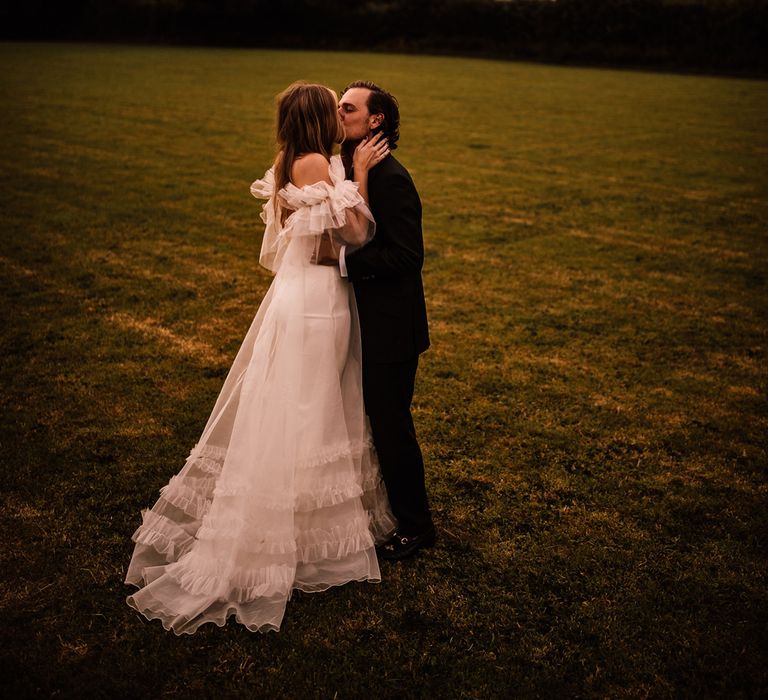 Bride in white Halfpenny London Mayfair dress with ruffle sleeves kisses groom in suit in field at garden wedding reception in Cornwall