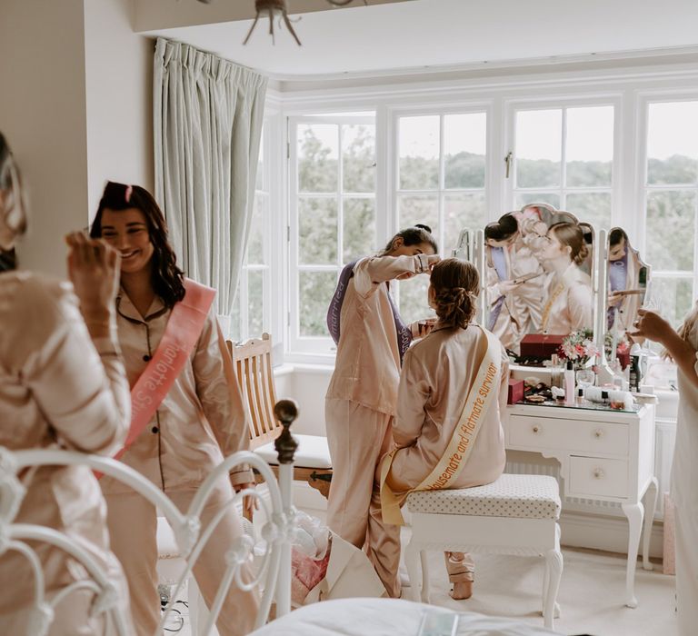 Bridesmaids in matching satin pyjamas and sashes have their make up done in bedroom before DIY garden wedding in Bedfordshire 