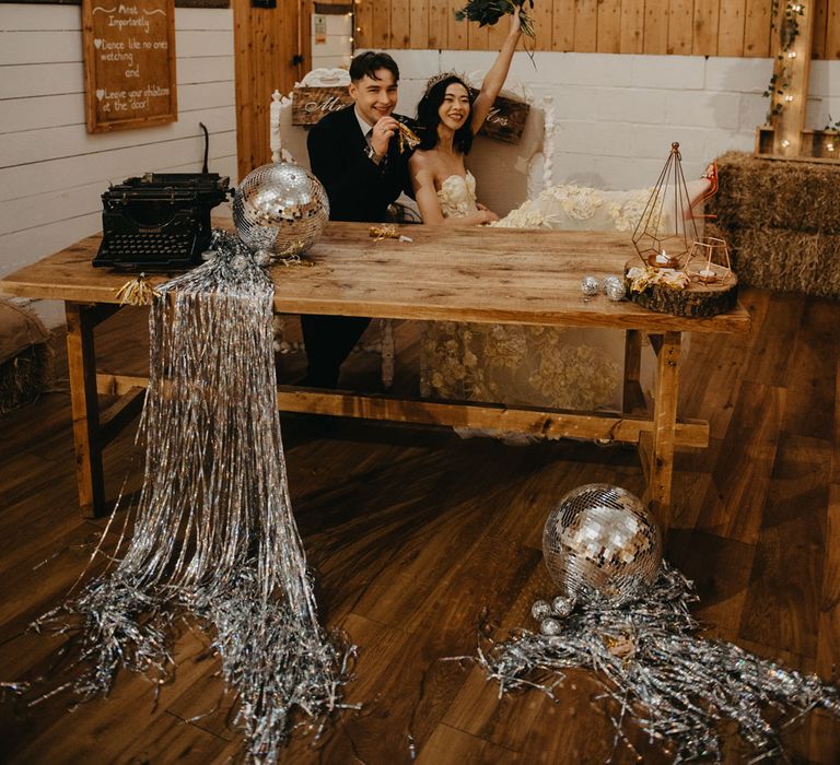 Bride in strapless feathered wedding dress and headpiece holds up bouquet as she sits with groom at wooden table for rustic wedding in Lancashire