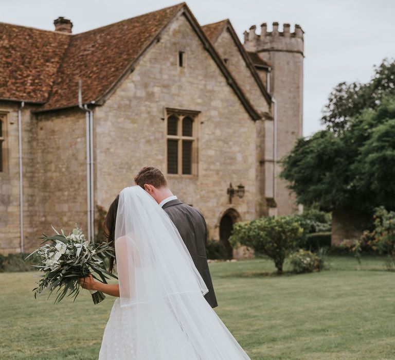 Bride in Pronovias wedding dress and cathedral veil walks through grounds of Notley Abbey holding white and green bridal bouquet with groom in grey suit