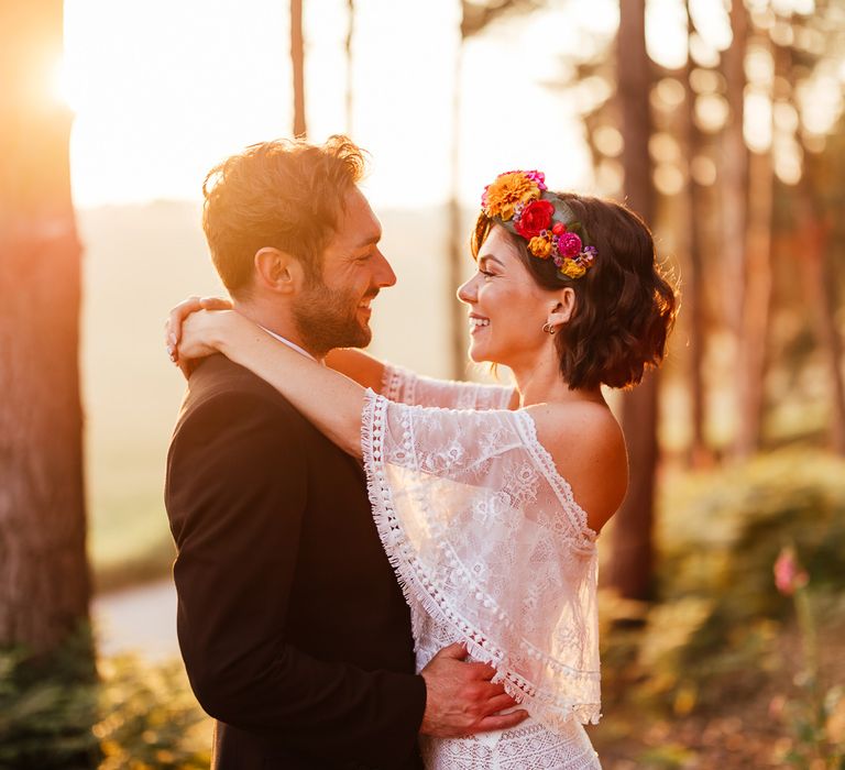 Smiling bride in off the shoulder Grace Loves Lace wedding Dres sand flower crown stands with smiling groom in black suit in woodland during golden hour