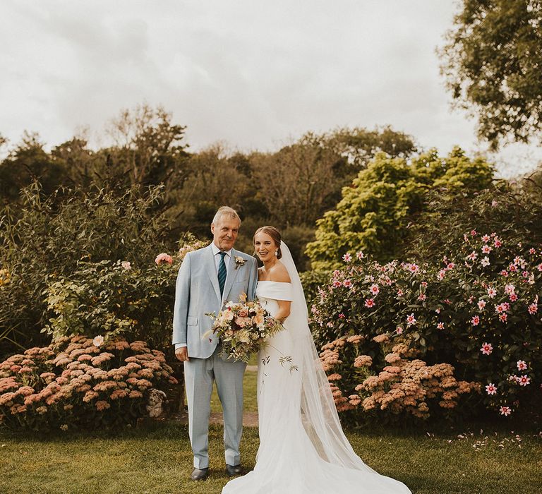 Bride stands with her father on her wedding day outdoors who wears pale blue suit