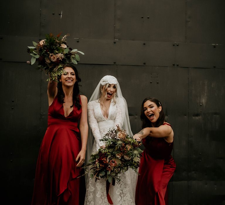 Boho bride in a lace wedding dress and Juliet cap veil standing with her bridesmaids in red satin bridesmaid dresses