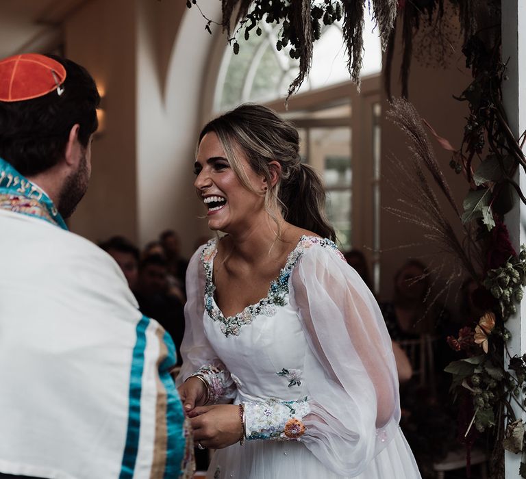 Laughing bride in mesh sleeve wedding dress with floral embroidery stands with groom in red kippah and tallit during wedding ceremony at Iscoyd Park