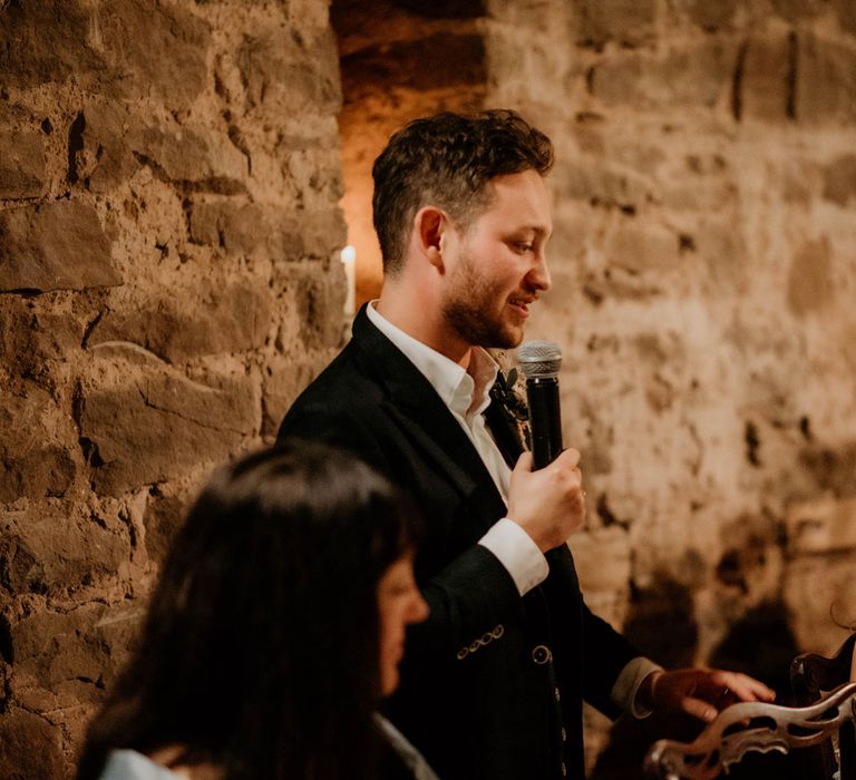 Bride in off the shoulder wedding dress looks at groom holding microphone as he makes speech during wedding reception