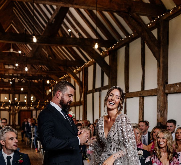 Groom in a navy suit holding hands with his bride in a silver sequin wedding dress at the altar of their rustic barn wedding 