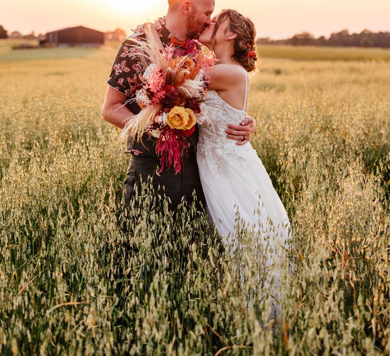 Bride in lace and tulle wedding dress kisses groom in patterned short sleeve shirt holding mixed rustic wedding bouquet as they stand in field during golden hour