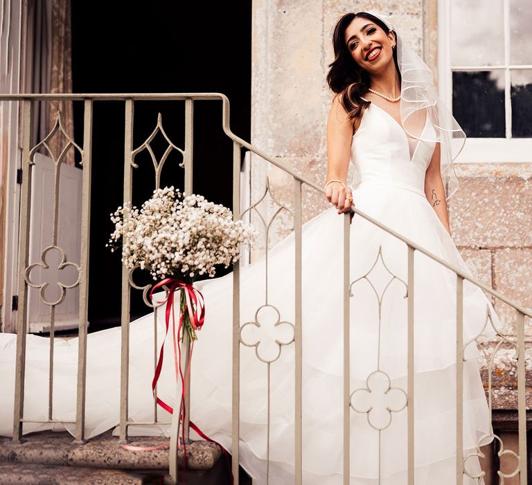 Smiling bride in headband, veil and layered tulle wedding dress stands on outdoor steps holding railing