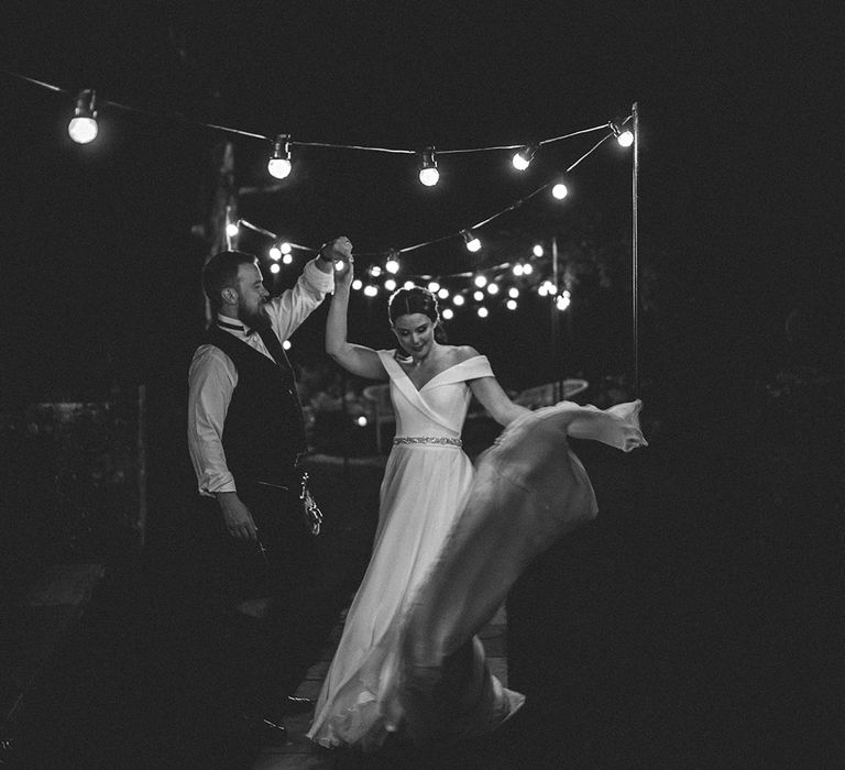 groom twirling his bride in a Suzanne Neville wedding dress under a canopy of festoon lights 