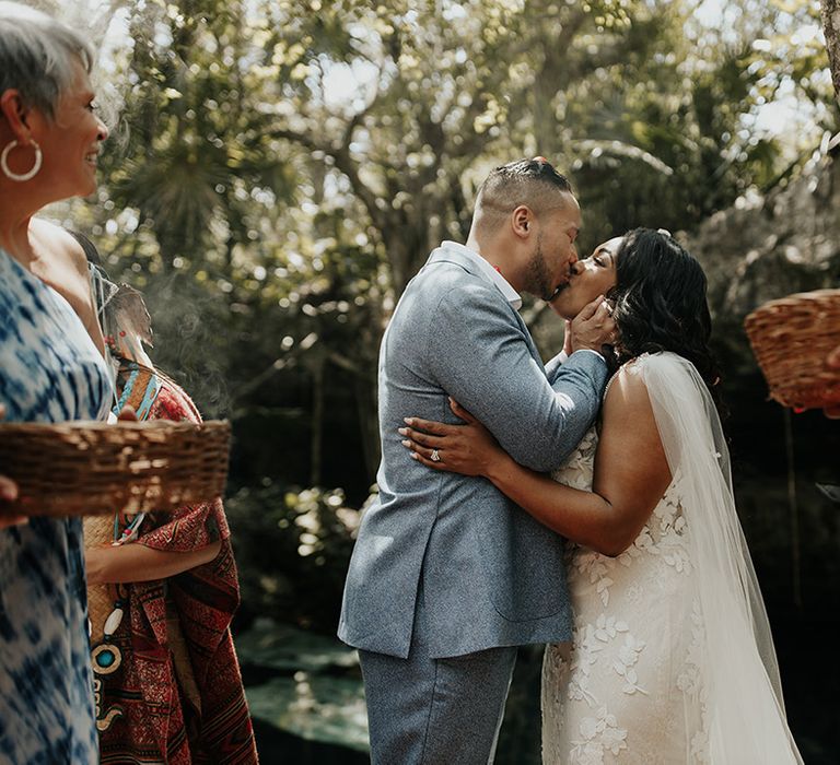 Groom in grey suit kisses bride in lace floral wedding dress in outdoor destination wedding