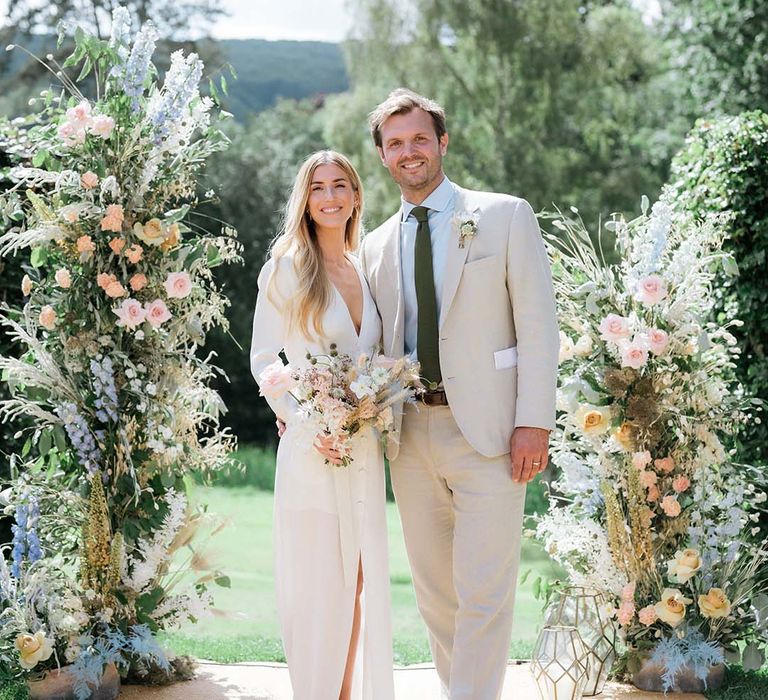 Bride in Bon Bride dress with plunging neckline with groom in cream suit in front of large pastel coloured flower displays