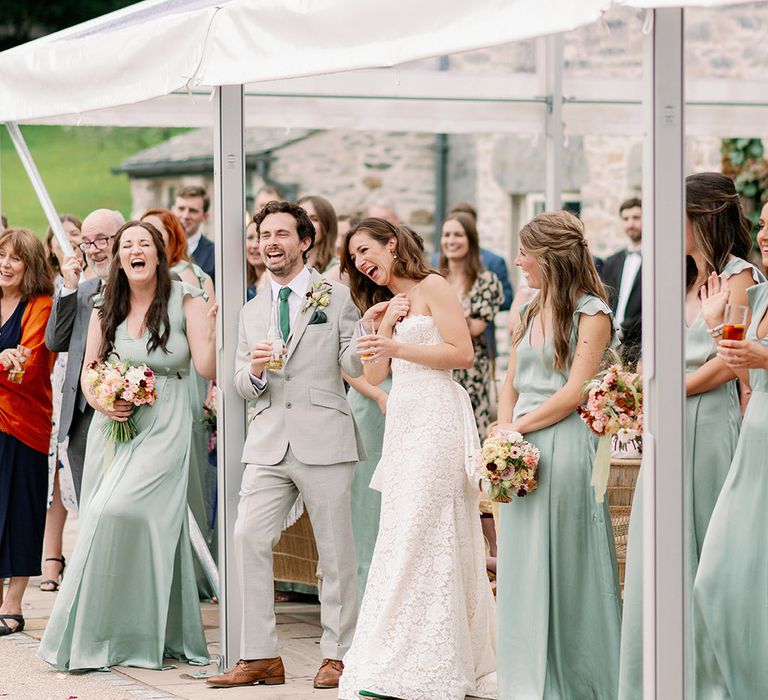 Bride and groom stand amongst their guests and bridesmaids in sage green dresses and laugh together