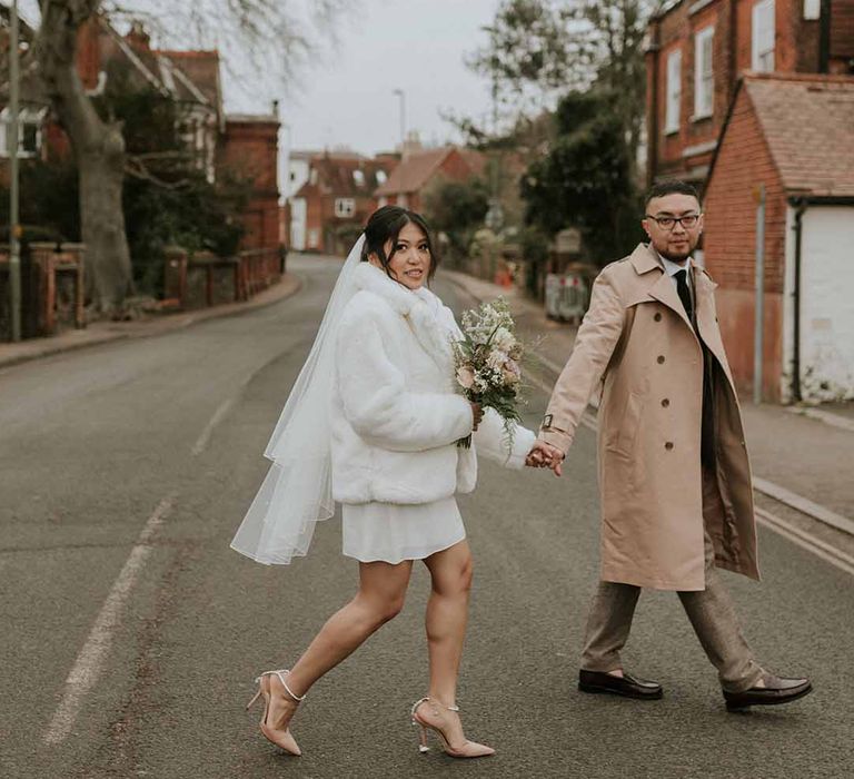 Bride in Vanessa Cocchiaro short wedding dress and white fur cropped coat and groom in Tru Clothing crossing the street