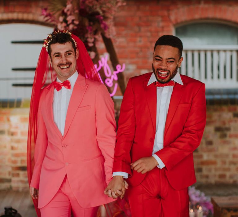 Groom in a pink suit, bow tie, gold crown and red veil laughing and holding hands with his partner in a red suit and bow tie at Prestwold Hall Barns 