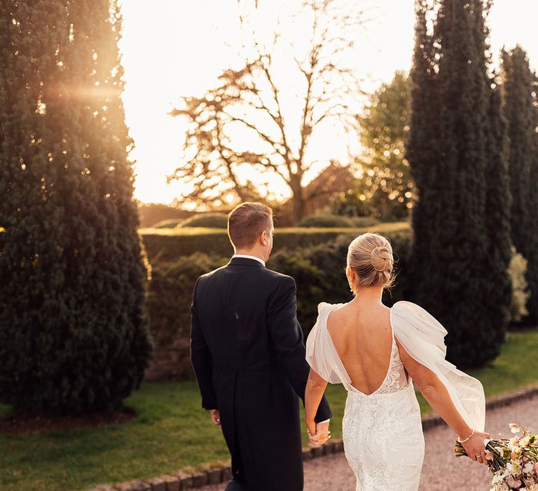 Bride in scoop back lace wedding dress with train holding pink wedding bouquet and hands with groom in morning suit 