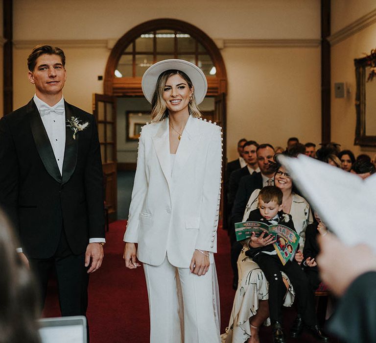 Bride and groom stand at the altar with groom in black tie with pale blue bow tie and bride in pearl trouser wedding suit 