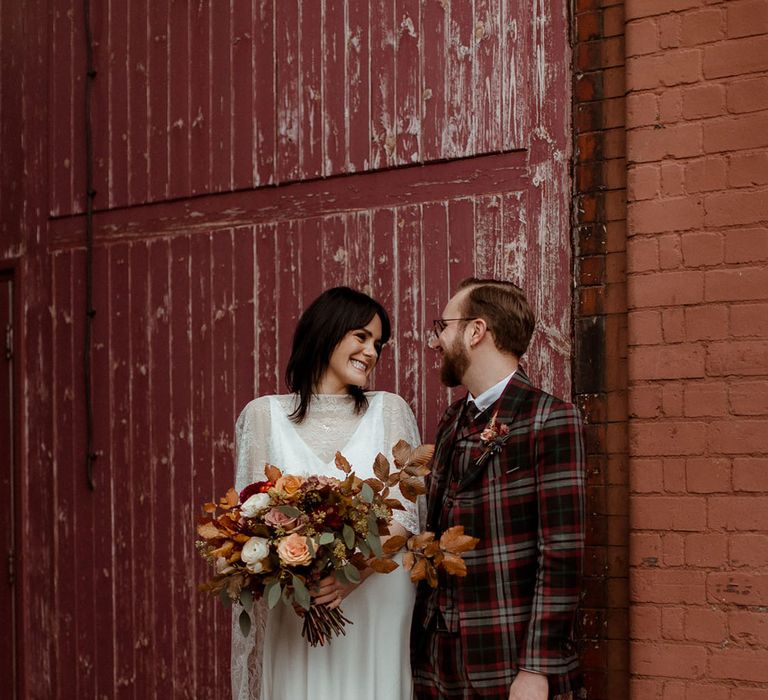 Bride & groom stand in front of industrial wall as bride holds Autumnal inspired bridal bouquet 