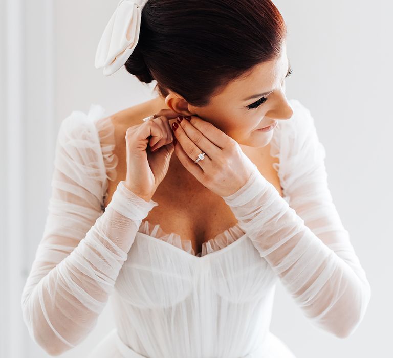 Bride in tulle ruffle detachable sleeve princess skirt wedding dress as she puts in her statement silver earrings with emerald cut engagement ring