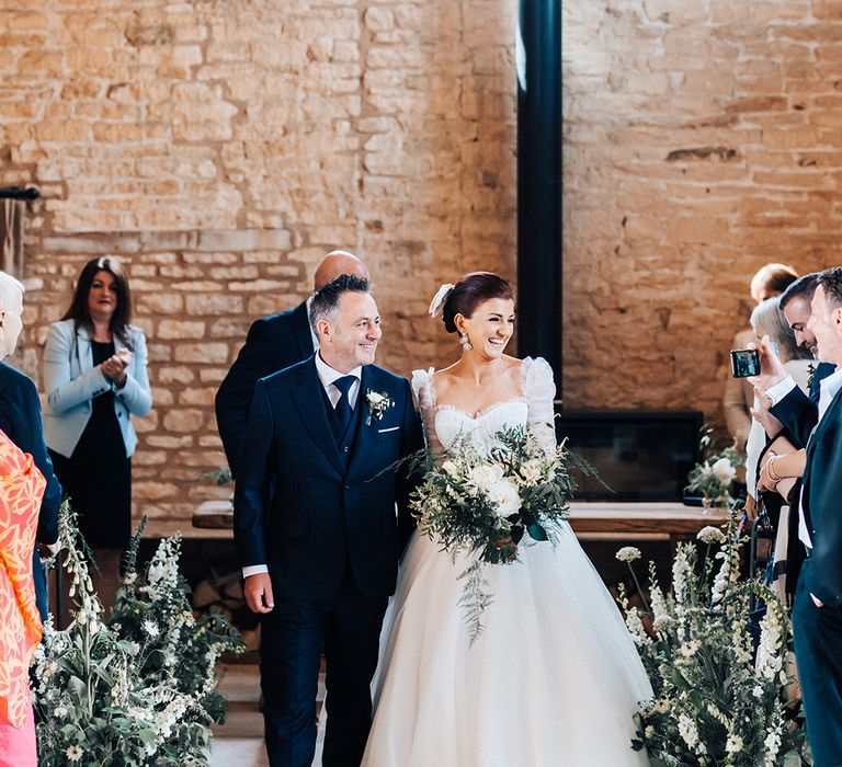 Groom in blue suit and bride in Emma Beaumont princess wedding dress walks back down the aisle as a married couple smiling to guests 