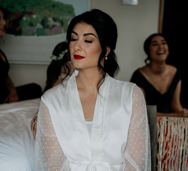 Bride in satin robe with sheer polka dot sleeves gets ready for her wedding day with red lipstick and black eyeliner 