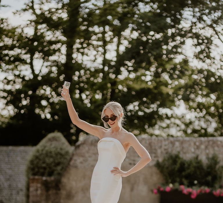 Bride in strapless wedding dress and cat eye black sunglasses raises her glass 