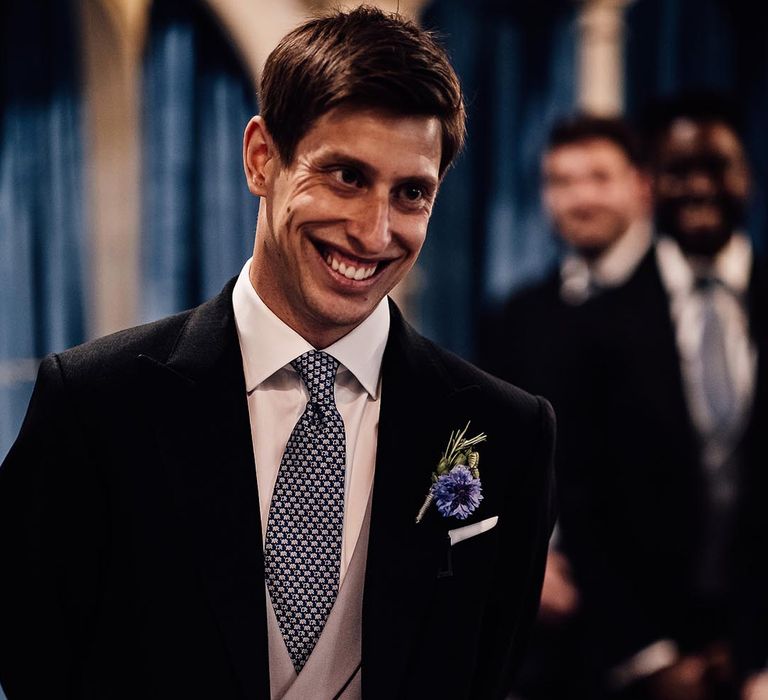 Groom smiles as he stands at the altar wearing a black suit with grey waistcoat and blue flower buttonhole 