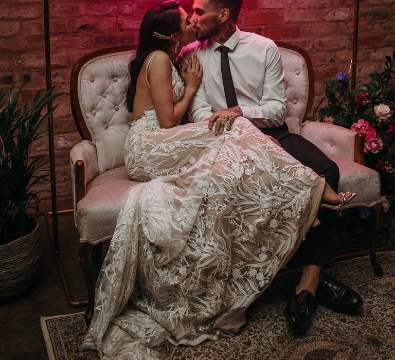 Bride and groom sit on a sofa and kiss under personalised pink neon sign 