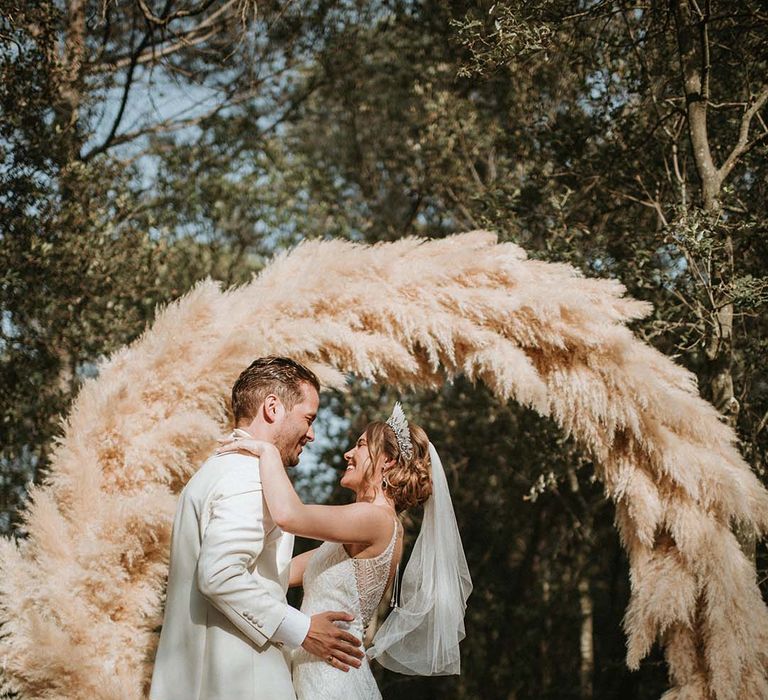 Bride and groom smile in front of their fluffy pampas grass moongate 