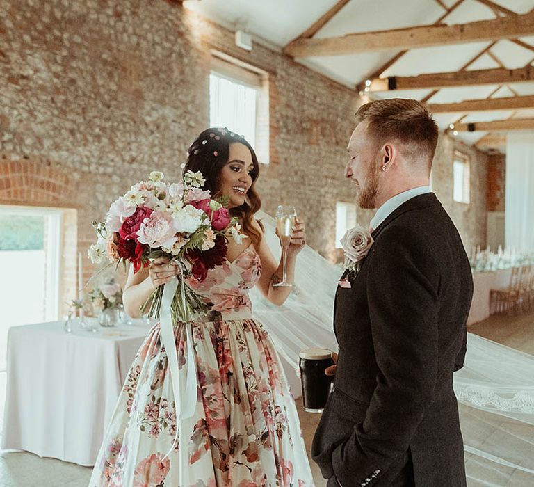 Bride in a Wendy Makin floral wedding dress holding a pink, white and red wedding bouquet with the groom in a suit drinking together 