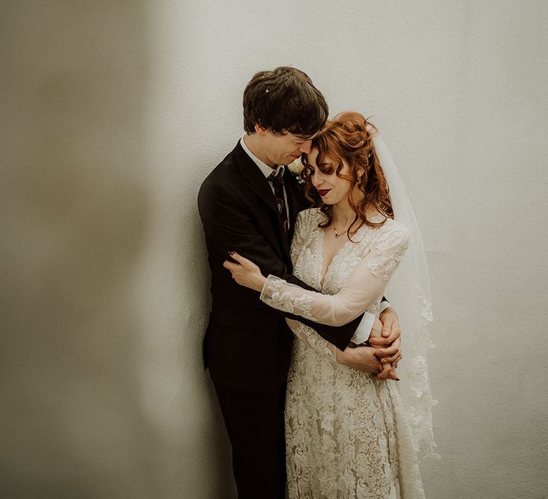 Bride wears long sleeve lace wedding dress with V-shaped neckline and embraces her groom in brown suit 