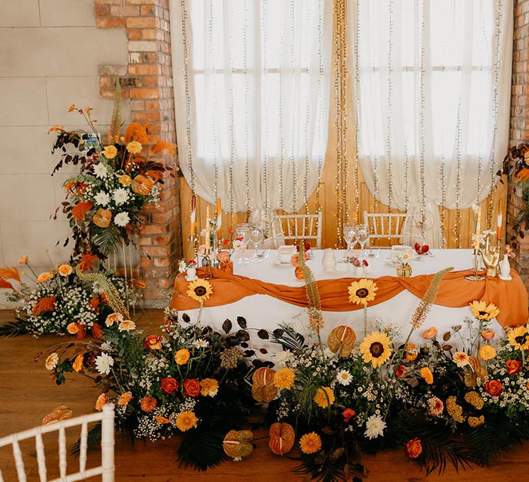 Sweetheart top table decorated with orange runner with orange and yellow wedding flowers including sunflowers, anthuriums and daisies