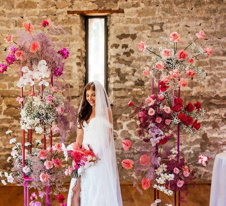 Bride wearing off-the-shoulder lace wedding dress holds bright pink floral bouquet and stands in front of bright floral wedding arch 