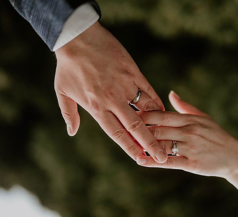 Bride and groom reach out and hold hands showing off their silver wedding rings and diamond engagement ring 