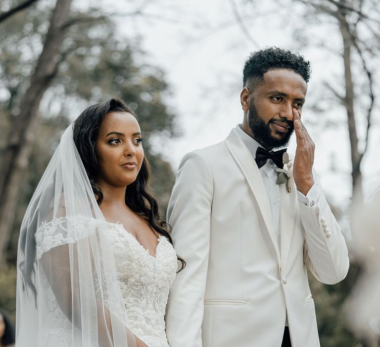 Bride wears off-the-shoulder lace wedding dress and cathedral veil as she holds the hand of her groom in black tie 