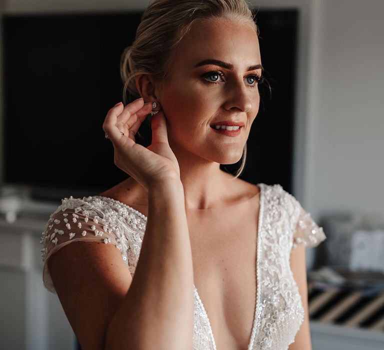 Bride places earrings in her ear as she wears her blonde hair pulled back into bun complete with pearl hair accessories 