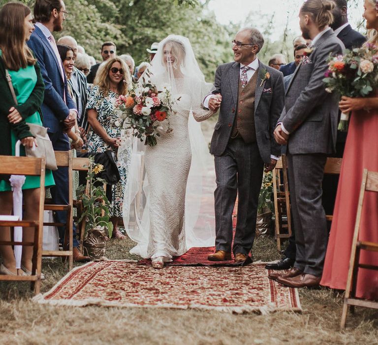 Father of the bride in a grey suit and brown waistcoat walks the bride in a lace wedding dress and veil down the aisle 