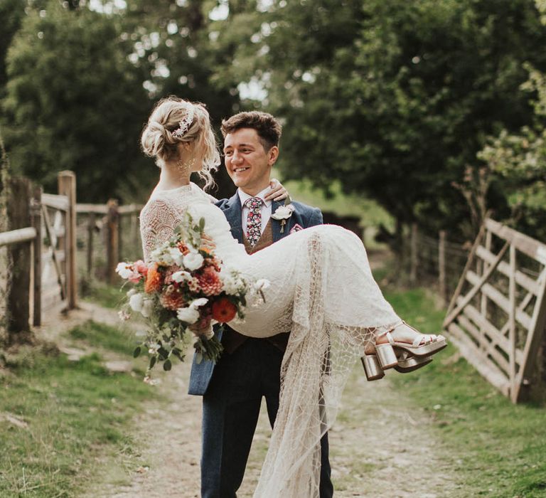 Bride in a long sleeve lace wedding dress with gold shoes being picked up by the groom in a blue suit 