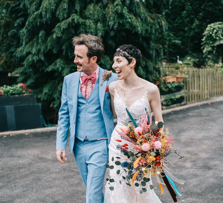 Bride wears fitted lace Enzoani wedding dress and carries bright floral bouquet alongside her groom