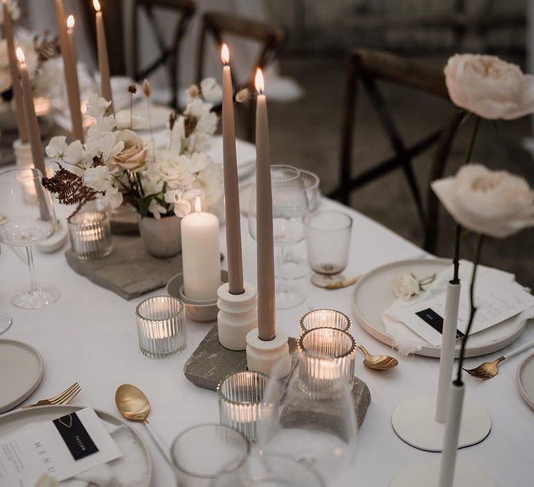 Neutral minimalistic wedding tablescape with long tapered beige candles, light grey ceramic plates and candle holders, neutral floral decorations and pillar candles