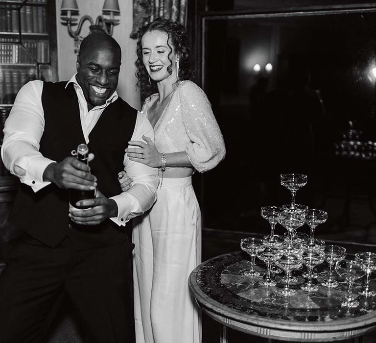 Groom in black waistcoat and suit trousers with bride in two piece outfit with sparkly long sleeve top and satin wide leg trousers popping champagne for champagne tower at Pepper Arden Hall