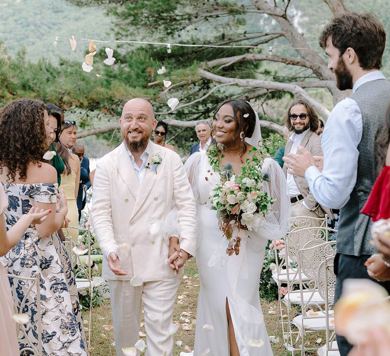 Bride and Groom walk through cream and peach coloured petal confetti whilst holding hands. The Brides something blue wedding shoes peep through her dress split