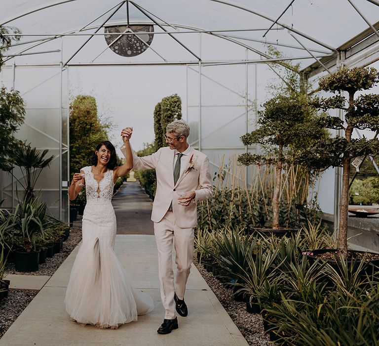 Bride and Groom walk through greenhouse holding hands in the air and smiling