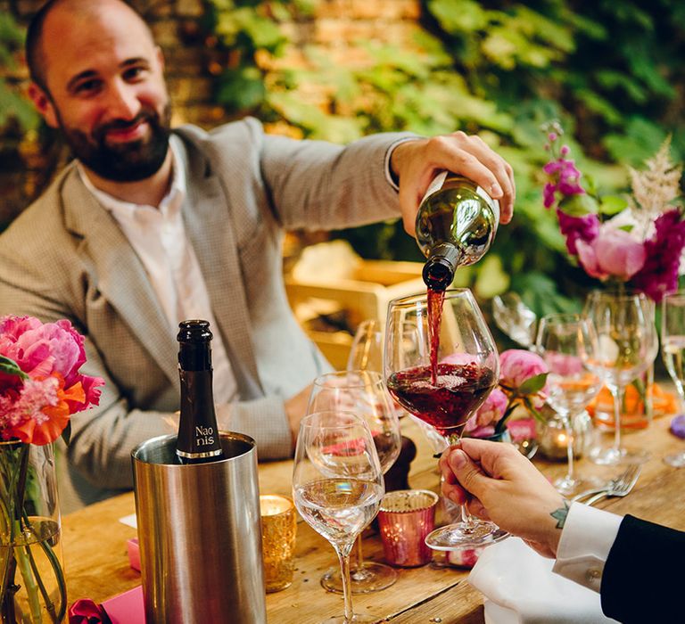 Wedding guests pouring a glass of red wine sitting at neutral, earthy wedding tablescape with pink and purple floral arrangements in long thin vases