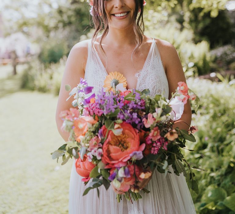 Bride wears scalloped lace wedding dress and colourful flower crown whilst holding matching bouquet 