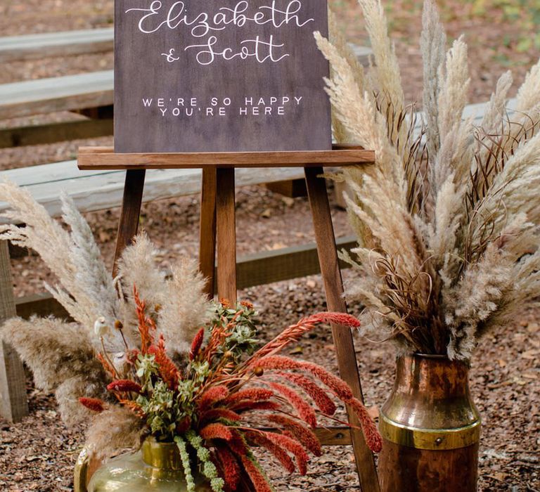 Hand made wood calligraphy wedding welcome sign with with white dried cotton stems, terra cotta celosia seeds and green foliage floral arrangements in large brass pots