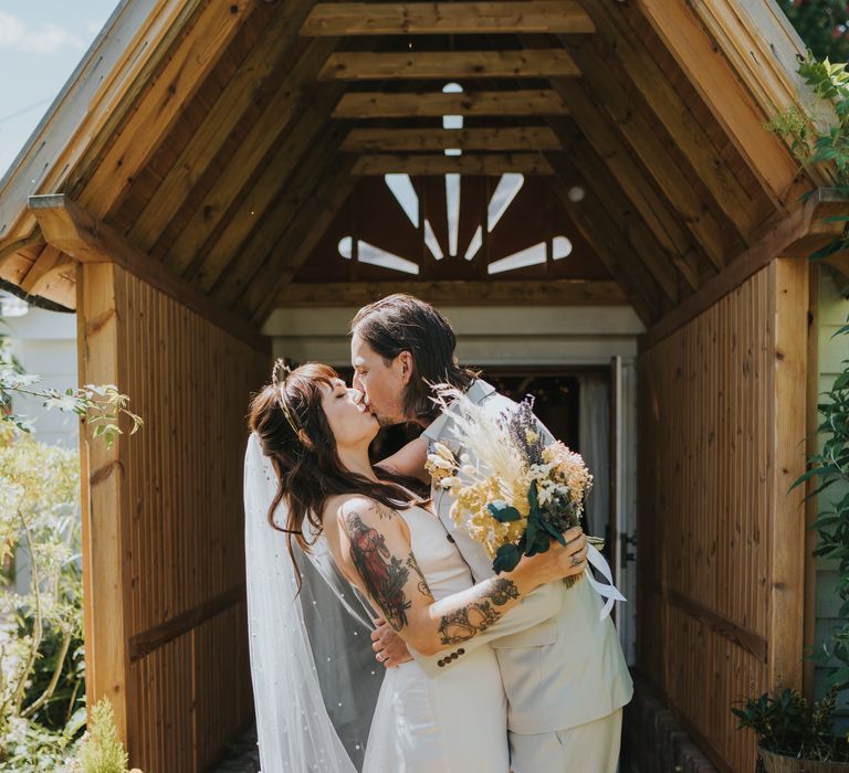 Bride in silk-look v-neck sleeveless ASOS wedding dress holding dried flower wedding bouquet with white bunny tails, green foliage, yellow dried gypsophila, lavender and baby’s-breath kissing groom in neutral coloured double-breasted ASOS suit and black high-top converse with dried flower and foliage boutonniere