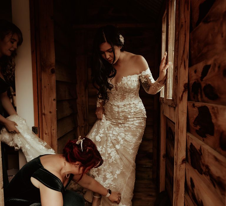 Bridesmaid helping bride get into her bridal outfit; off the shoulder bodycon wedding dress with long, sheer sleeves and lace detailing and strappy open toe wedding heels with large polka dot bow 